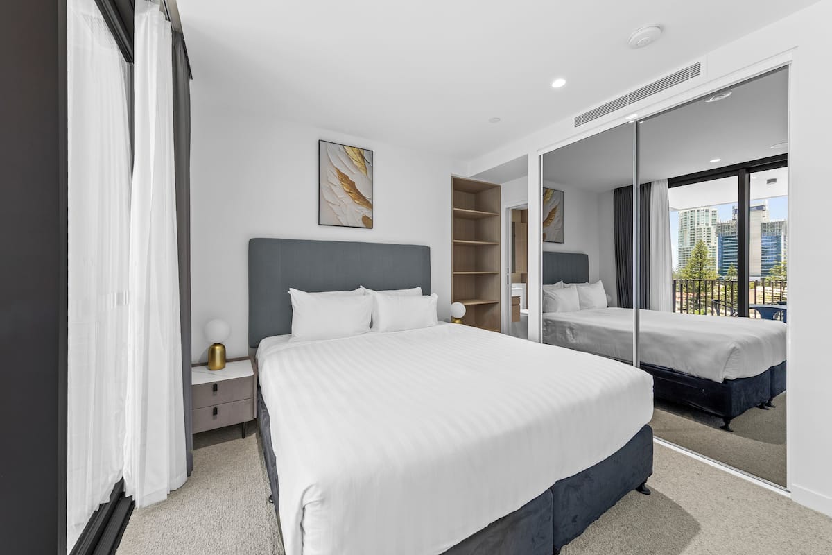 Una habitación de hotel moderna con una cama grande, una mesita de noche, un espejo y vistas a la ciudad desde la ventana.