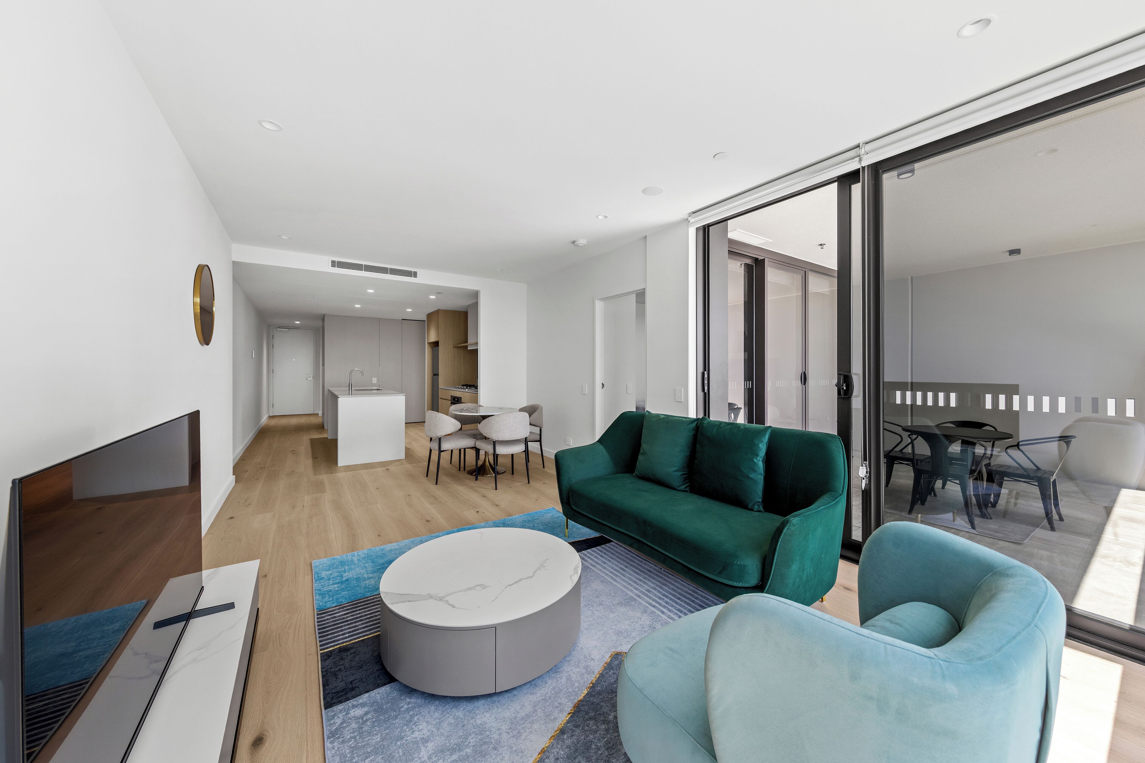 A modern living room with a green sofa, a blue armchair, and a round coffee table. There is a flat-screen TV on a stand, a dining area with a table and chairs, and a kitchen in the background.