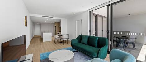 A modern living room with a green sofa, a blue armchair, and a round coffee table. There is a flat-screen TV on a stand, a dining area with a table and chairs, and a kitchen in the background.