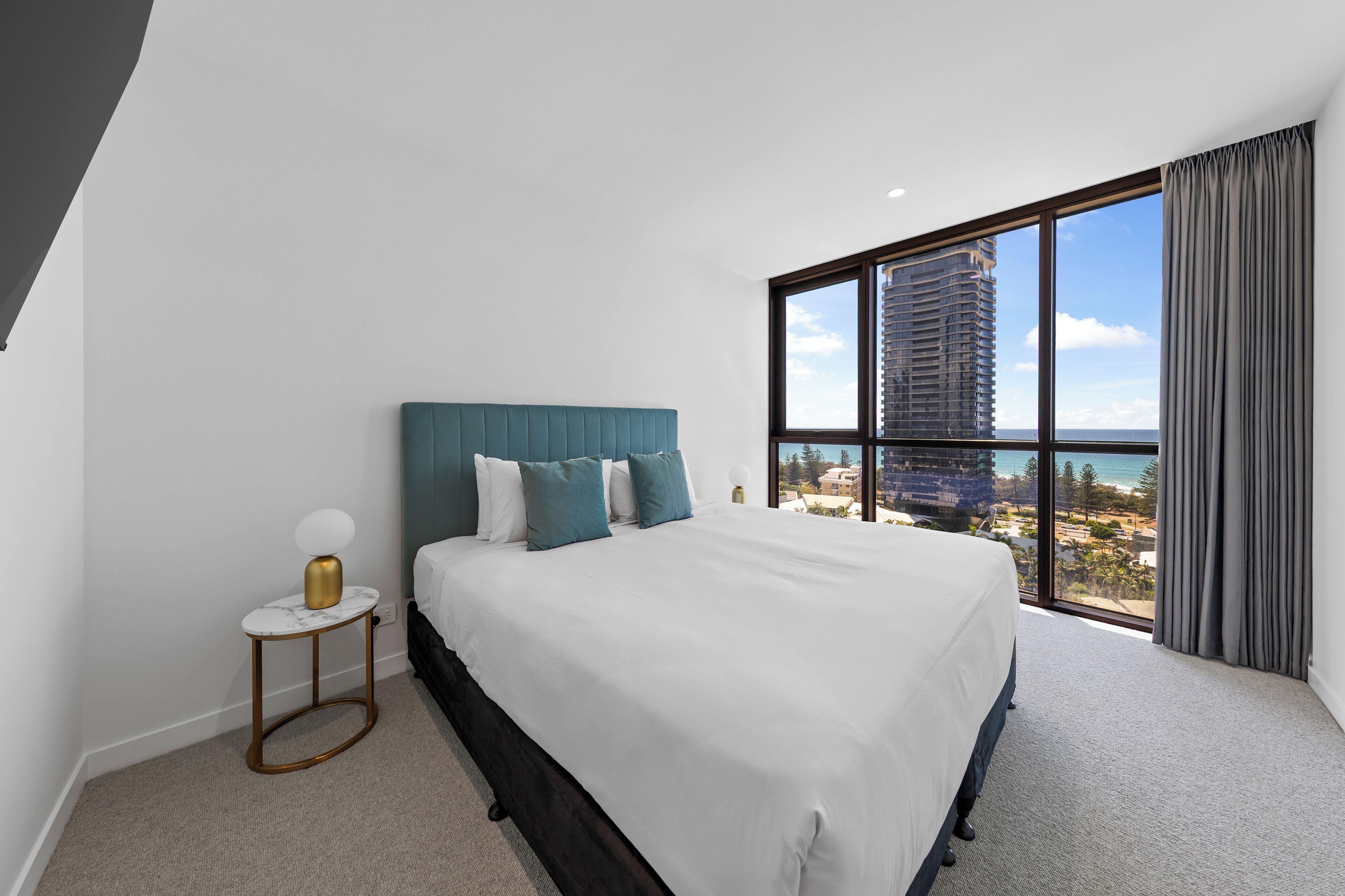 Habitación de hotel moderna con una cama grande, una mesita auxiliar y vistas a un paisaje urbano a través de un ventanal amplio.