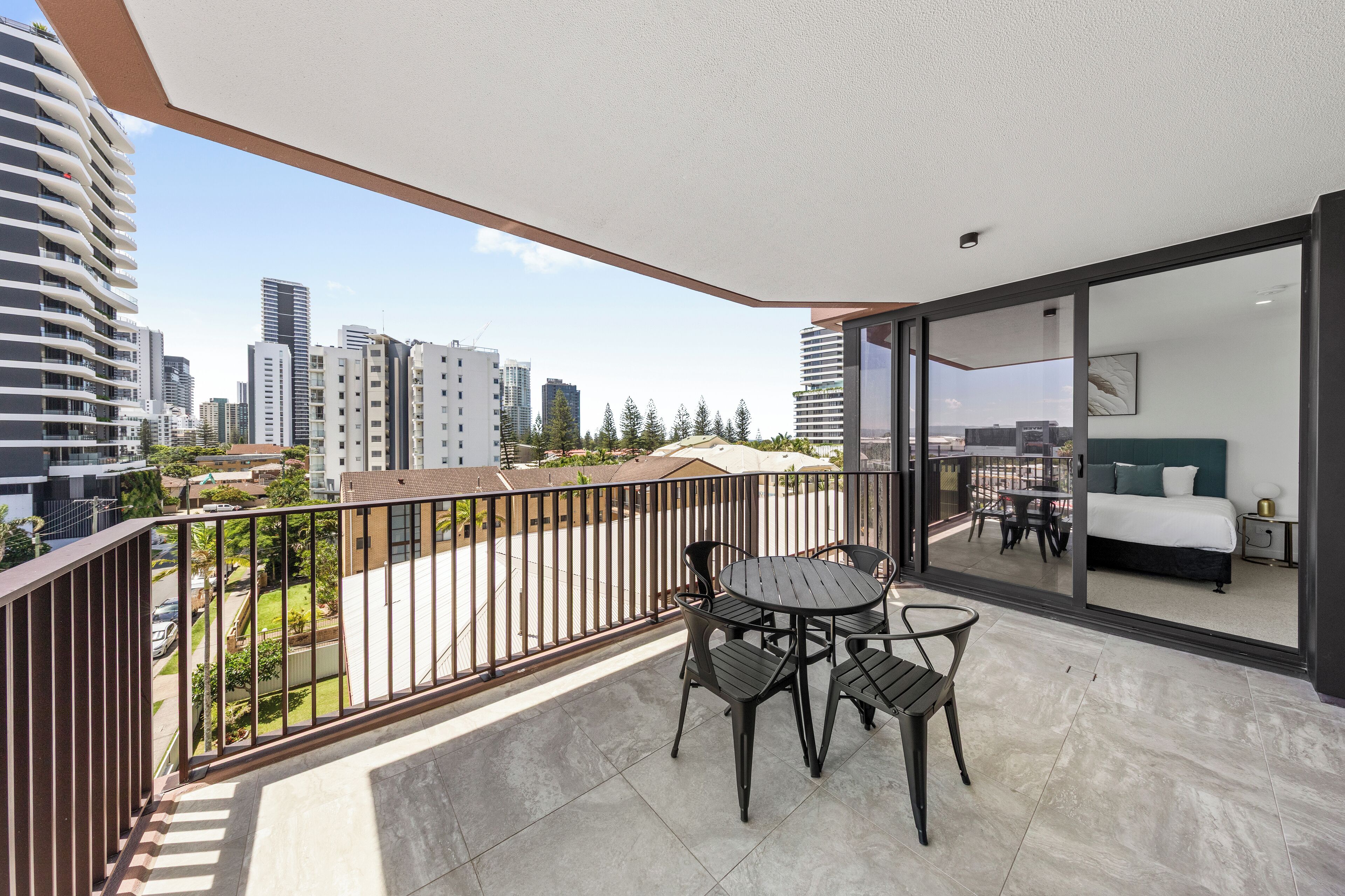 A balcony with a table and chairs, a bed with a green headboard, and a city view.