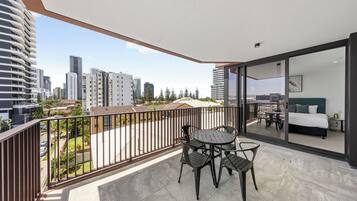 A balcony with a table and chairs, a bed with a green headboard, and a city view.