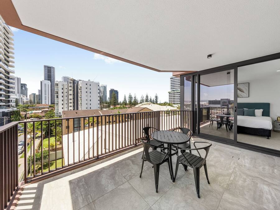 A balcony with a table and chairs, a bed with a green headboard, and a city view.