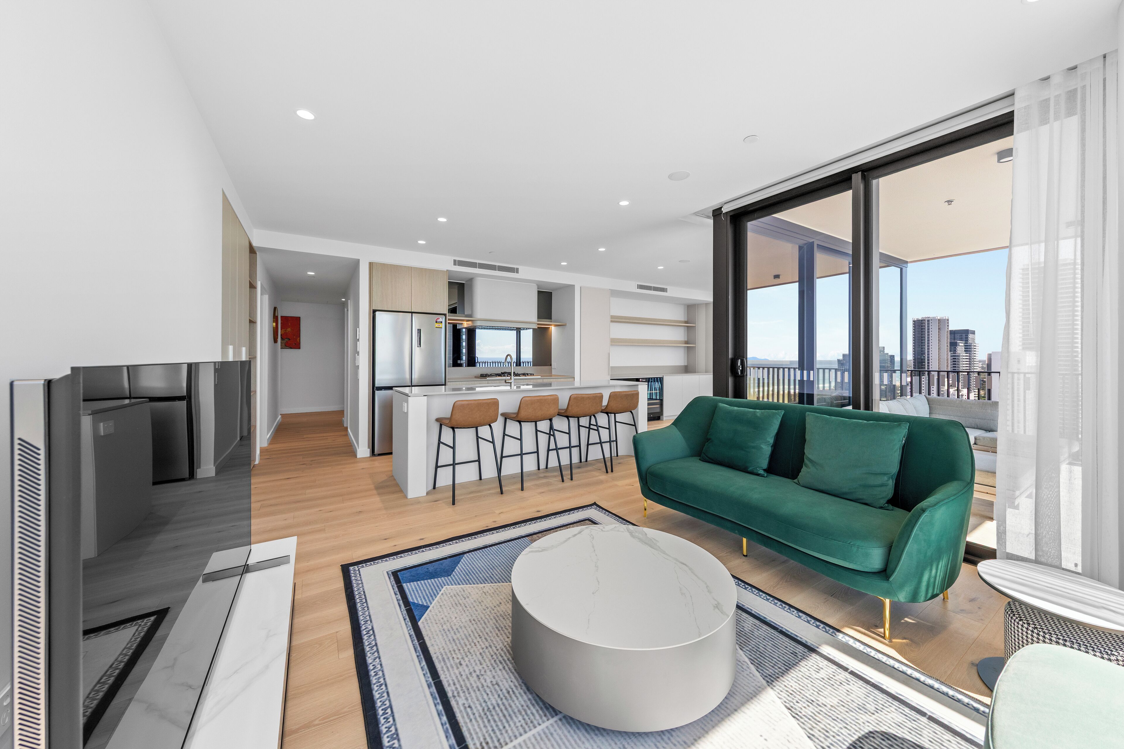 Ein modernes Wohnzimmer mit einem grünen Sofa, einem runden Couchtisch und einem Fernseher. Es gibt eine Küchenzeile mit einer Bar und Hockern. Das Zimmer verfügt über große Fenster mit Blick auf die Stadt.