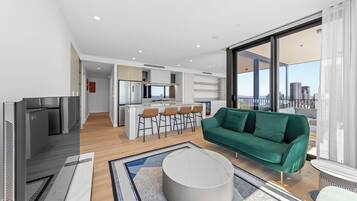 Ein modernes Wohnzimmer mit einem grünen Sofa, einem runden Couchtisch und einem Fernseher. Es gibt eine Küchenzeile mit einer Bar und Hockern. Das Zimmer verfügt über große Fenster mit Blick auf die Stadt.