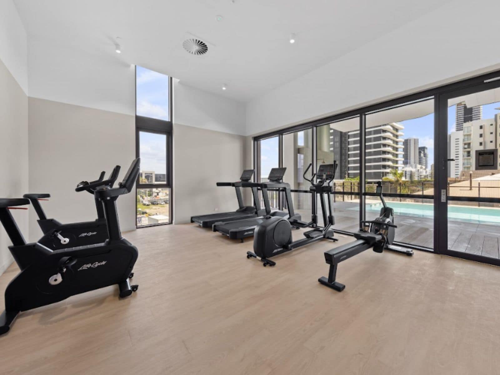 A modern gym with cardio machines, a treadmill, and a rowing machine, featuring large windows and a view of the city.