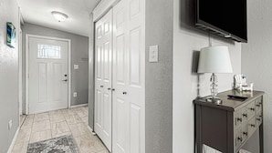 A hallway with a white door, a set of double doors, a television mounted on the wall, and a small table with a lamp and remote control.