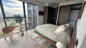 A modern bedroom with a large bed, a chair, a TV, and a view of the city through large windows. - High-floor apartment with city views (Guadalajara)