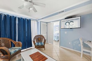 Quarto com ventilador de teto, televisão exibindo 'Ocean City Vacation Homes', mobiliário de vime e vista para outro cômodo.