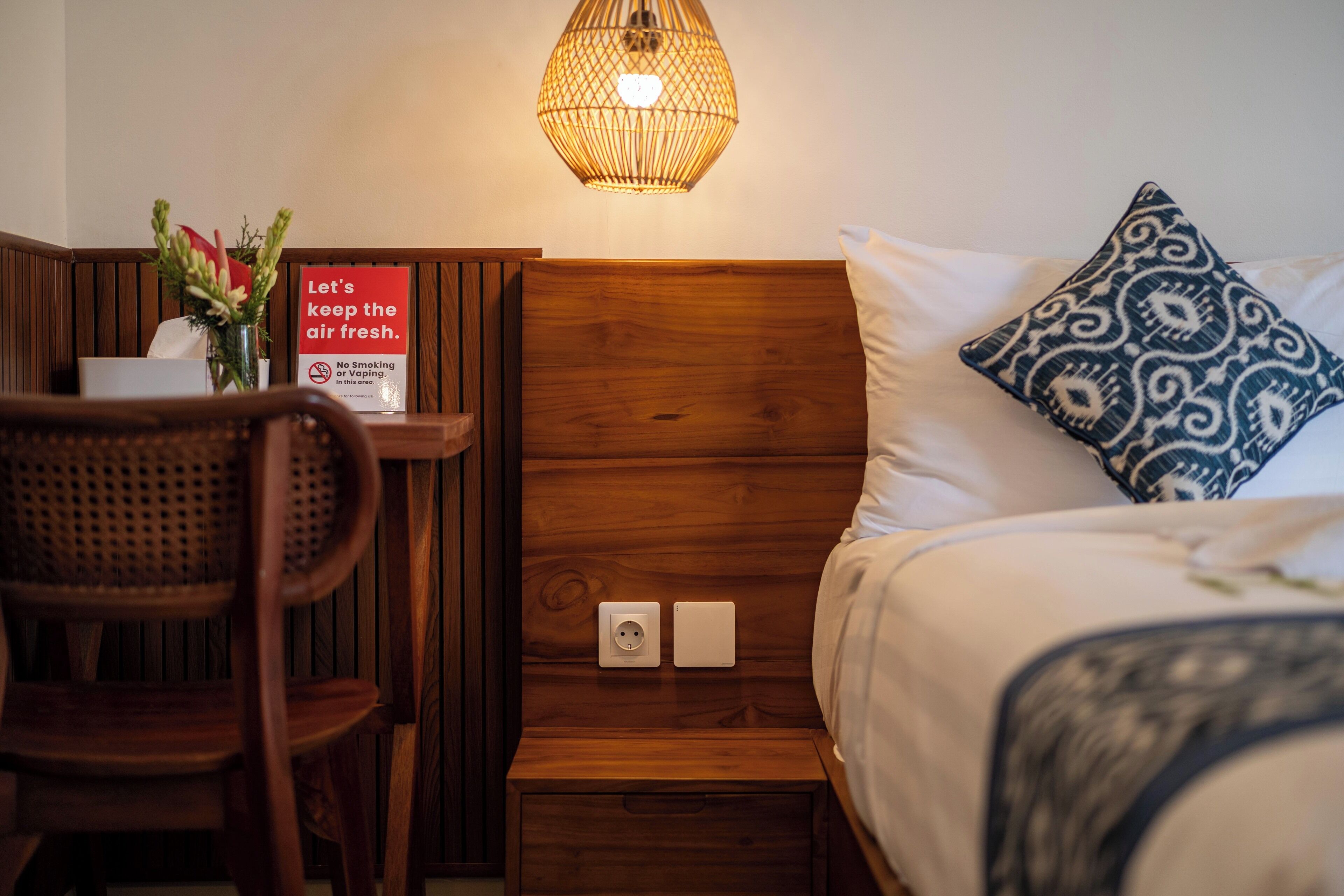 Habitación de hotel con cabecera de madera, mesita de noche con un letrero sobre frescor del aire, una silla y un cojín estampado.