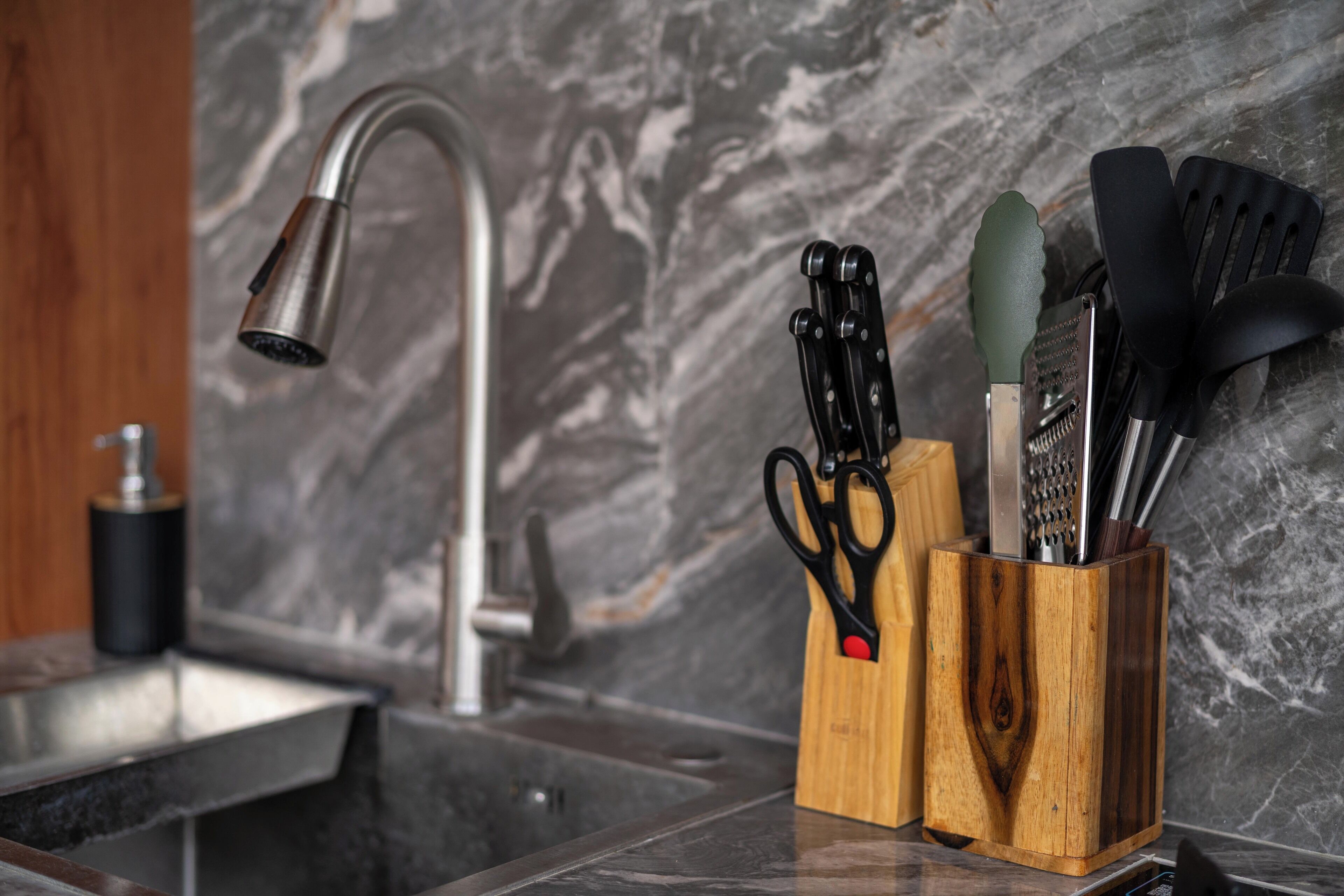 A kitchen with a marble backsplash, a modern faucet, and a wooden utensil holder with scissors, a grater, and a spatula.
