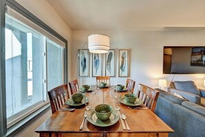 A dining area with a wooden table set for six, surrounded by chairs, with a large window on one side and a flat-screen TV mounted on the wall opposite. - Downtown 2BD 1BA - 2 Blocks to Main/River #187 (Buena Vista)