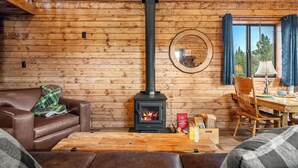 Een gezellig chaletinterieur met een houtkachel, een leren bank, een eettafel en uitzicht op de natuur door een raam.