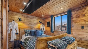 Een houten slaapkamer met een bed, twee kussens, een bankje met een plaid en twee jassen hangen aan de muur.
