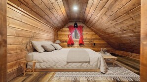 Um quarto de sótão acolhedor, com paredes de madeira, uma cama com almofadas, uma mesinha de cabeceira, uma mesa e uma janela com cortinas vermelhas.