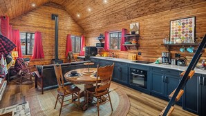Interior acolhedor de cabana com teto de madeira, área de jantar com mesa redonda e cadeiras, cozinha com armários azuis e uma lareira.
