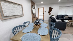 A dining area with a wooden table, blue chairs, a decorative centerpiece, and a living room with a sofa and a coffee table. - Clematis Cove • Private Hot Tub + Fenced Yard • Near Downtown & Siesta Key (Sarasota)