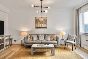 Ein modernes Wohnzimmer mit einem grauen Sofa, zwei Beistelltischen mit Lampen, einem Couchtisch und einem Fernseher auf einem Standfuß.