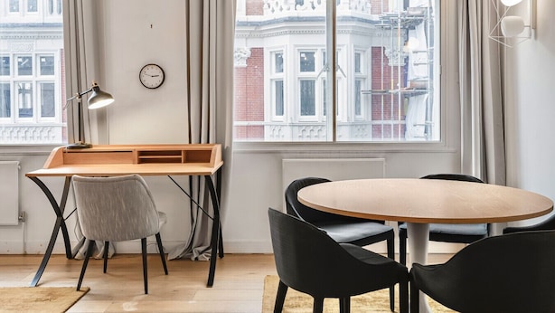 A modern room with a desk, chairs, a round table, a clock, and a view of buildings through large windows. - Mayfair 2-bed With Private Parking Near Buckingham Palace Green Park (London)