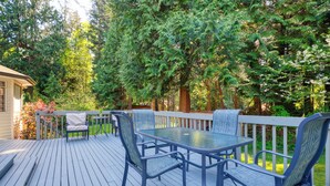 Une terrasse en bois avec une table et des chaises en verre, entourée d’une végétation luxuriante et d’arbres.