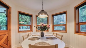 Une salle à manger avec une table ronde, des chaises en bois, des sets de table en fibres tressées, un vase de fleurs et de grandes fenêtres donnant sur un paysage arboré.