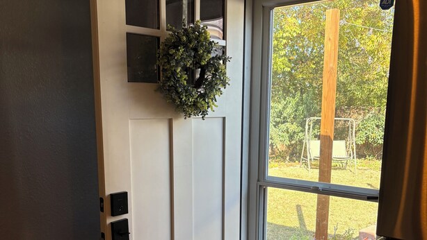 Eine weiße Tür mit einem kleinen Blumentopf obenauf, ein Fenster mit Blick auf einen Garten und ein Klingel.