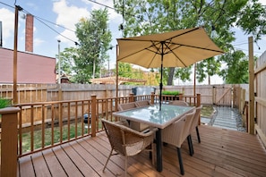A wooden deck with a glass-top table and wicker chairs, an umbrella, and a wooden fence.