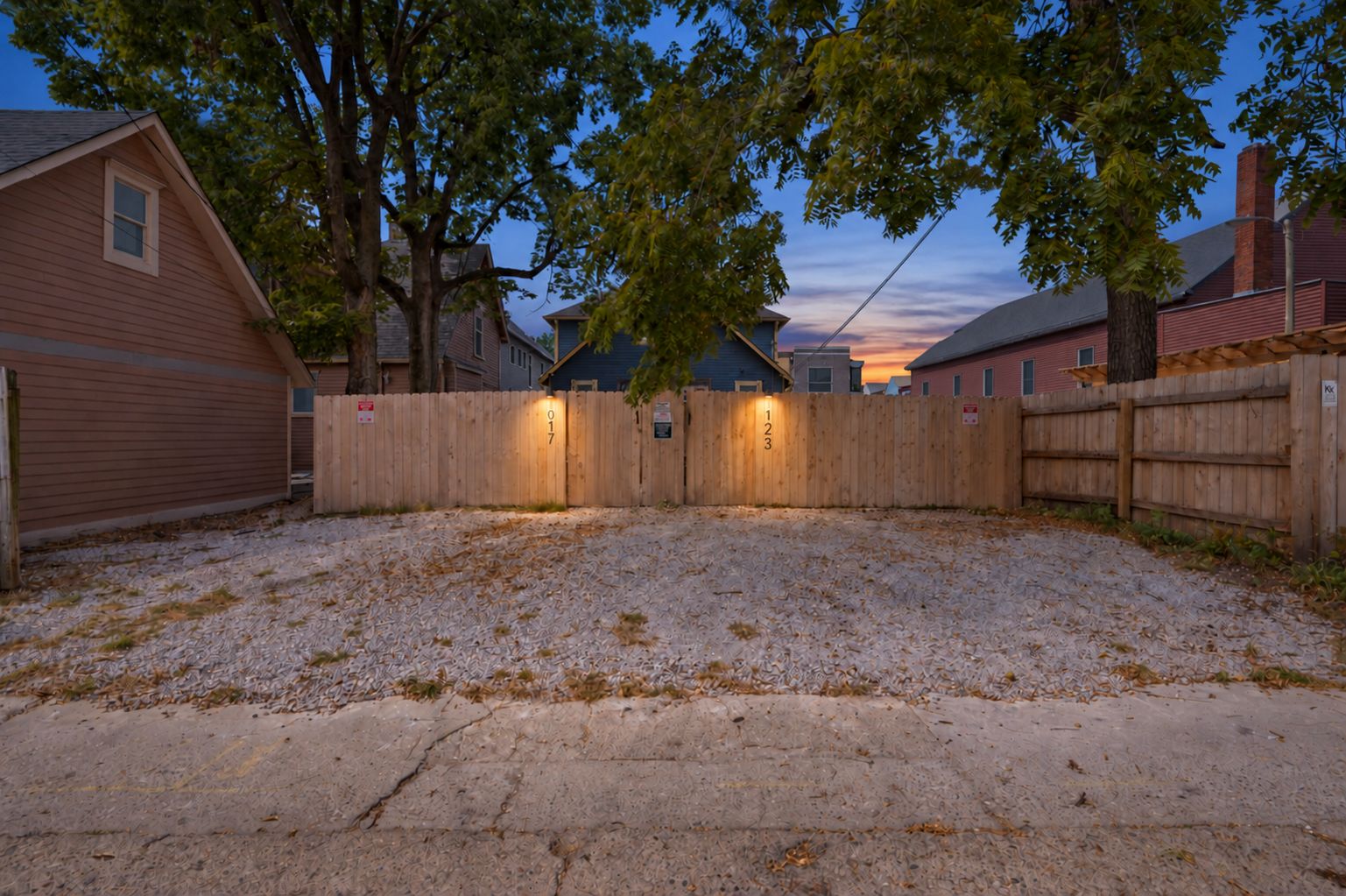 A gravel driveway leading to a wooden fence with numbered gates, surrounded by residential buildings.