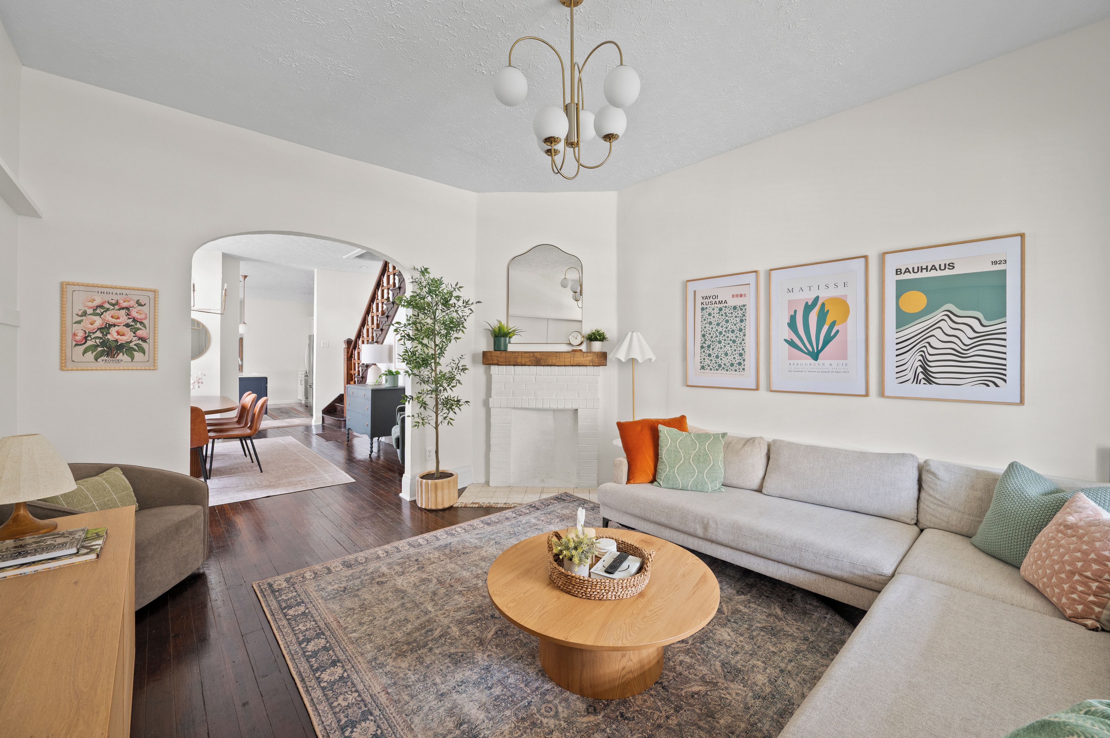 A modern living room with a sofa, coffee table, and a dining area with chairs. There are framed artworks on the walls and a staircase leading to another level.