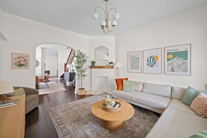 A modern living room with a sofa, coffee table, and a dining area with chairs. There are framed artworks on the walls and a staircase leading to another level. - Walk to Mass Ave: HOT TUB, private yard, parking (Indianapolis)