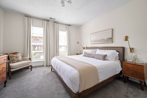 A bedroom with a large bed, a nightstand, a chair, a bedside table, and a window with curtains.