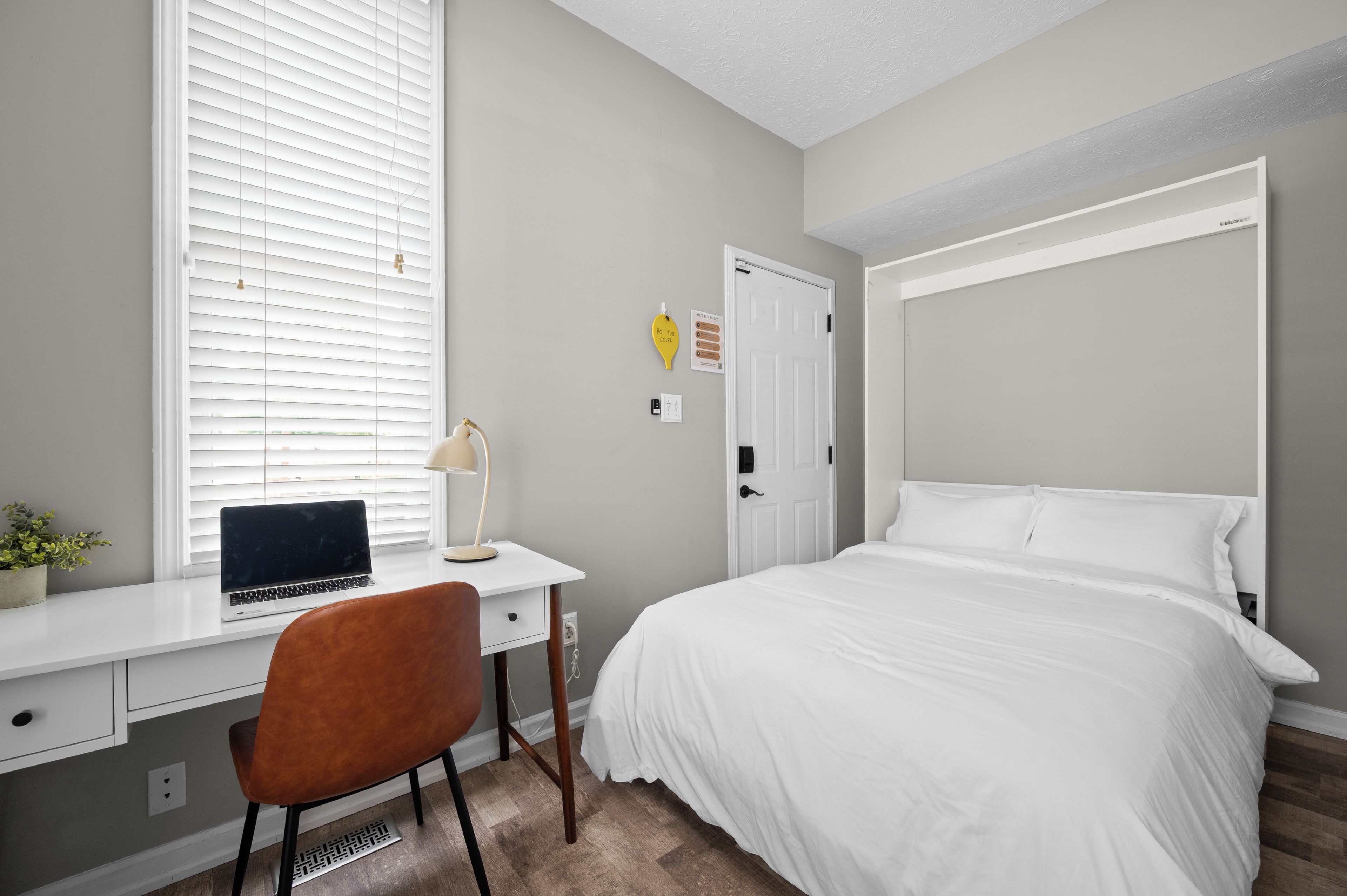 A bedroom with a bed, a desk with a laptop, a chair, and a window with blinds.