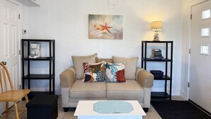 Ein Wohnzimmer mit einem beigefarbenen Sofa, einem Couchtisch, einem Holzstuhl und zwei schwarzen Regalen mit Büchern und Dekoration.