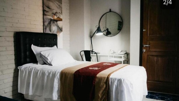 Ein Einzelbett mit Kopfteil, ein Nachttisch mit Lampe, ein runder Spiegel an der Wand und eine Holztür.