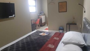 Ein Hotelzimmer mit Bett, Schreibtisch, Stuhl, einem kleinen Tisch, einem an der Wand montierten Fernseher und einem gerahmten Schild an der Wand.