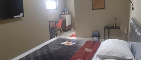 A hotel room with a bed, a desk, a chair, a small table, a wall-mounted TV, and a framed sign on the wall.