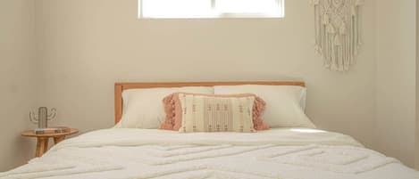 A bedroom with a bed, a window, a macrame wall hanging, and a small table with a cactus decoration.