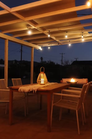 Een terras buiten met een houten tafel en stoelen, een hanglamp en lichtslinger.