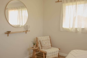 A room with a round mirror, a wooden chair with a cushion, a small table with a plant, and a window with sheer curtains.