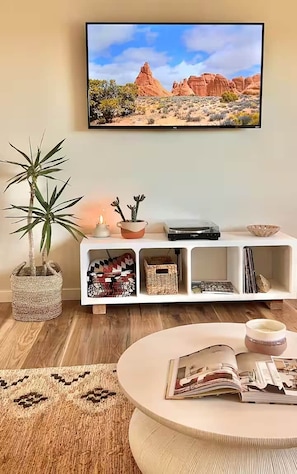 A living room with a flat-screen TV mounted on the wall, a white console table with decorative items, a round coffee table with magazines, and a potted plant.