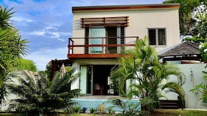 Ein modernes, zweistöckiges Haus mit Balkon, Swimmingpool und üppiger tropischer Vegetation.