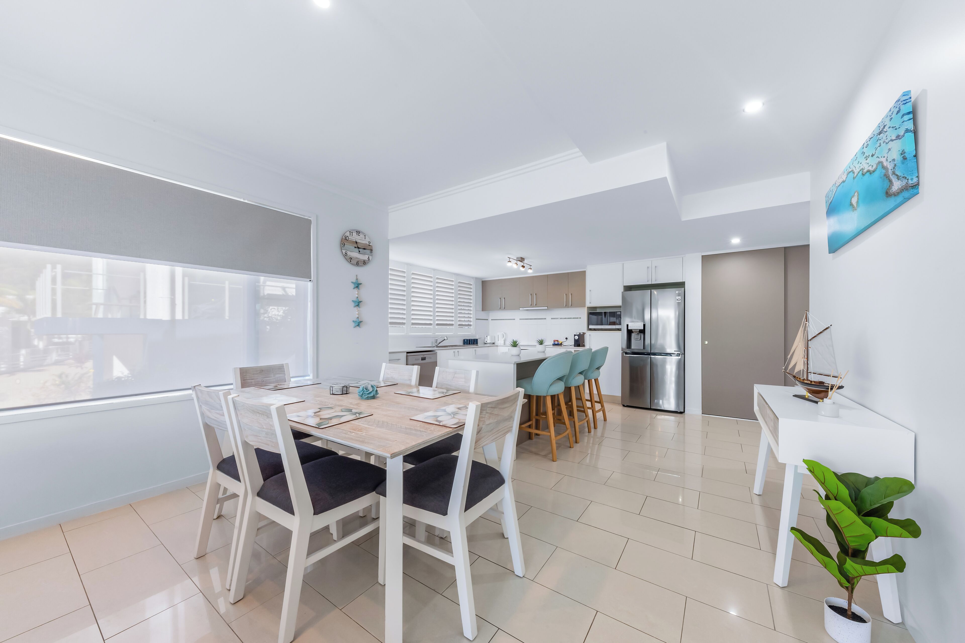 Una cocina moderna con armarios blancos, un refrigerador de acero inoxidable y una zona de comedor con mesa y sillas de madera.