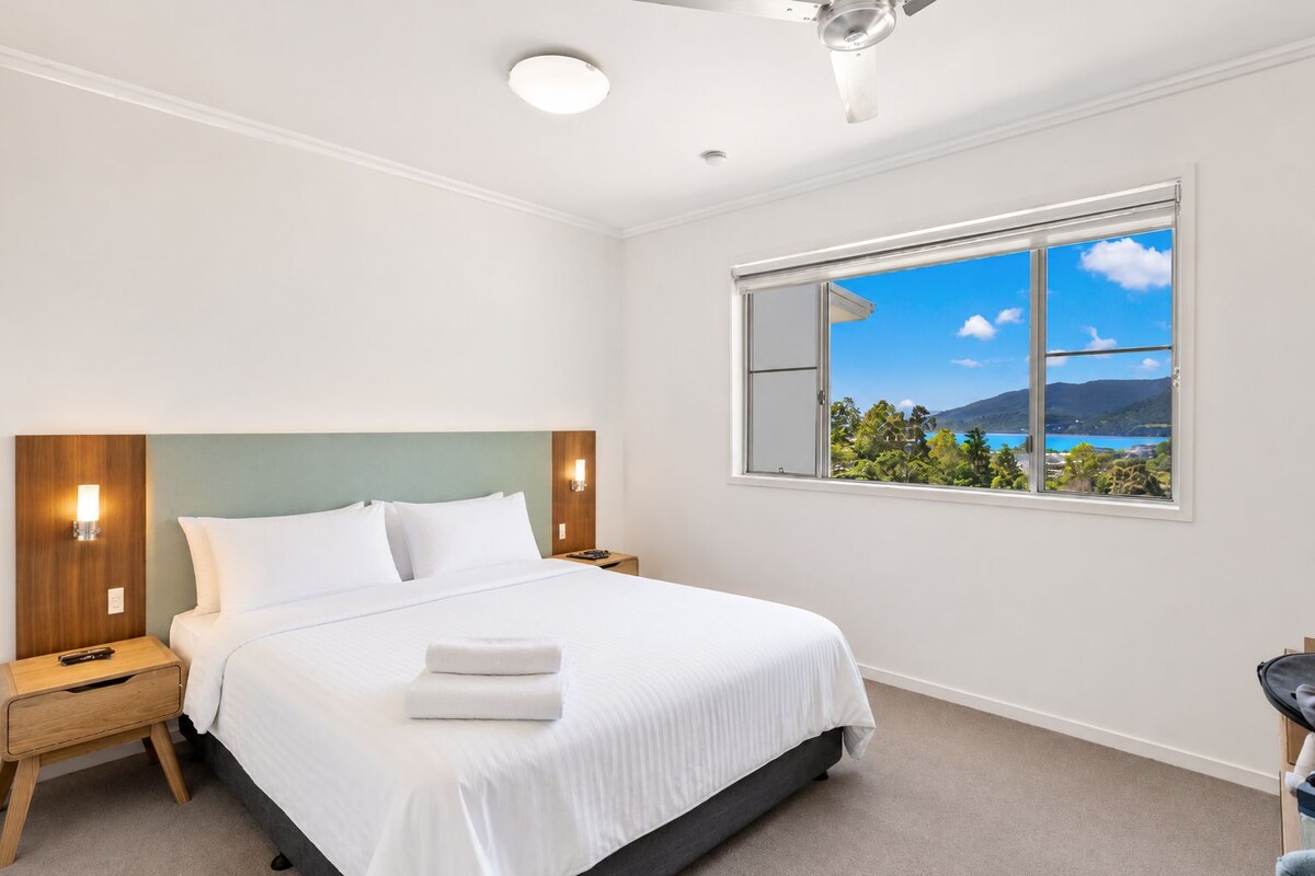 A hotel room with a bed, nightstands, a ceiling fan, a window with a view of a landscape, and a wall-mounted light fixture.