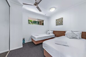 A hotel room with a large bed, a smaller bed, a ceiling fan, a window with a view of greenery, and a wall-mounted artwork. - Summit Apartments Airlie Beach (Airlie Beach)