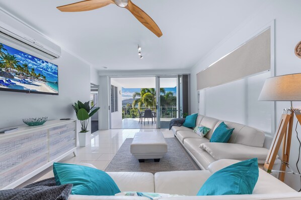 A modern living room with a ceiling fan, a large flat-screen TV, a sofa with teal cushions, and a view of a balcony with a glass door.