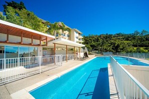 Una piscina con valla blanca, sillas de descanso y un edificio con balcones y una zona cubierta.