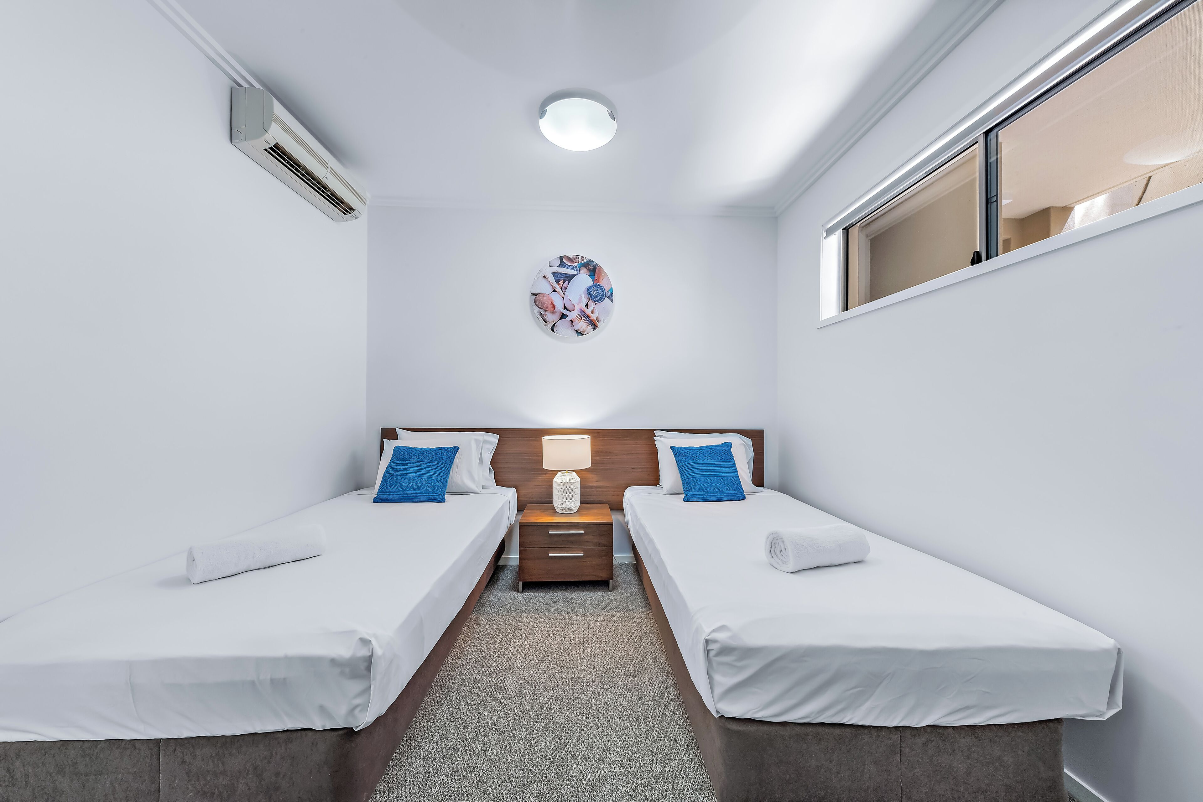 A hotel room with two single beds, a nightstand, a lamp, a wall-mounted air conditioner, and a ceiling light.