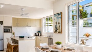 A modern kitchen with white cabinets, a central island, and a dining area with white chairs and a wooden table. - Summit Apartments Airlie Beach (Airlie Beach)