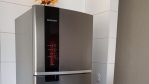 A stainless steel refrigerator with a digital control panel, placed in a room with white tiled walls and a red storage bin nearby. - Beach House (Praia Grande)