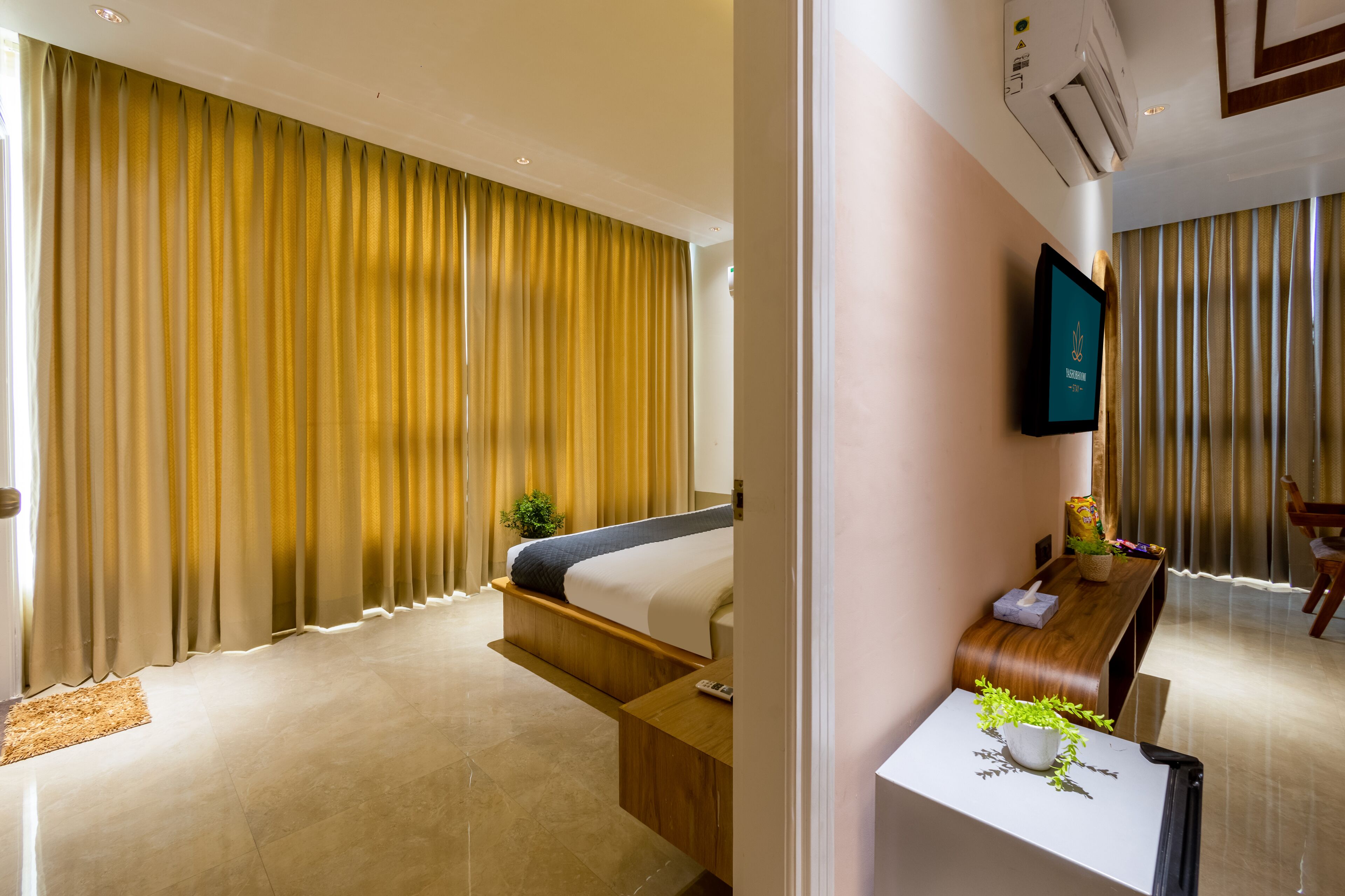 A modern hotel room with a large bed, a wooden bench, a small table with a plant, and a TV mounted on the wall.